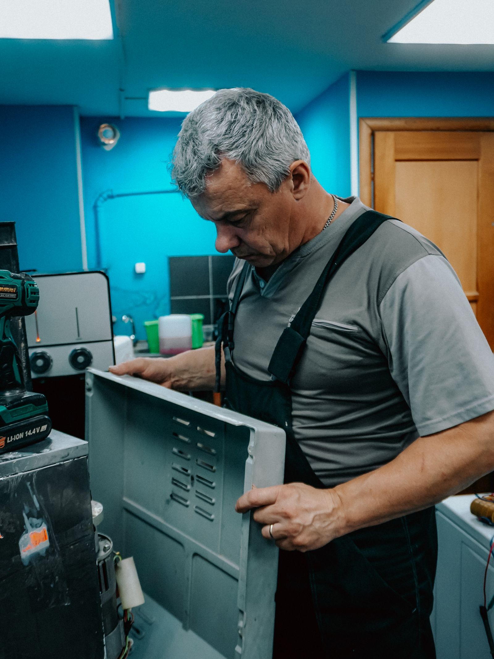 A grey-haired technician in grey overalls in a workshop with teal-blue walls removing the sheet-metal back panel from a large domestic appliance, a cordless drill resting on top of the machine.