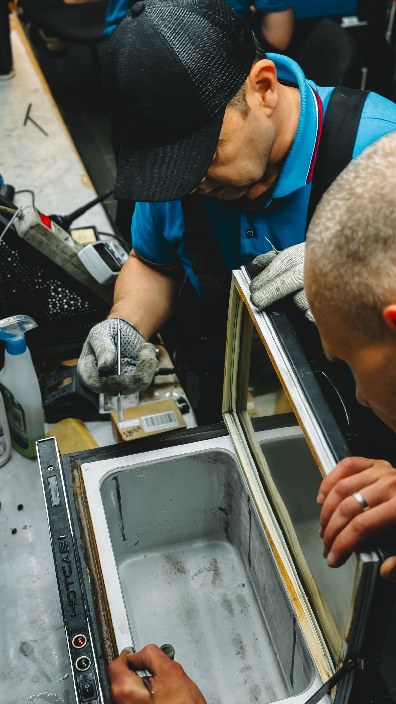 Two technicians in blue polo shirts and grey work gloves working together on a stainless-steel commercial chest freezer with its lid propped open, a spray bottle and hand tools nearby.