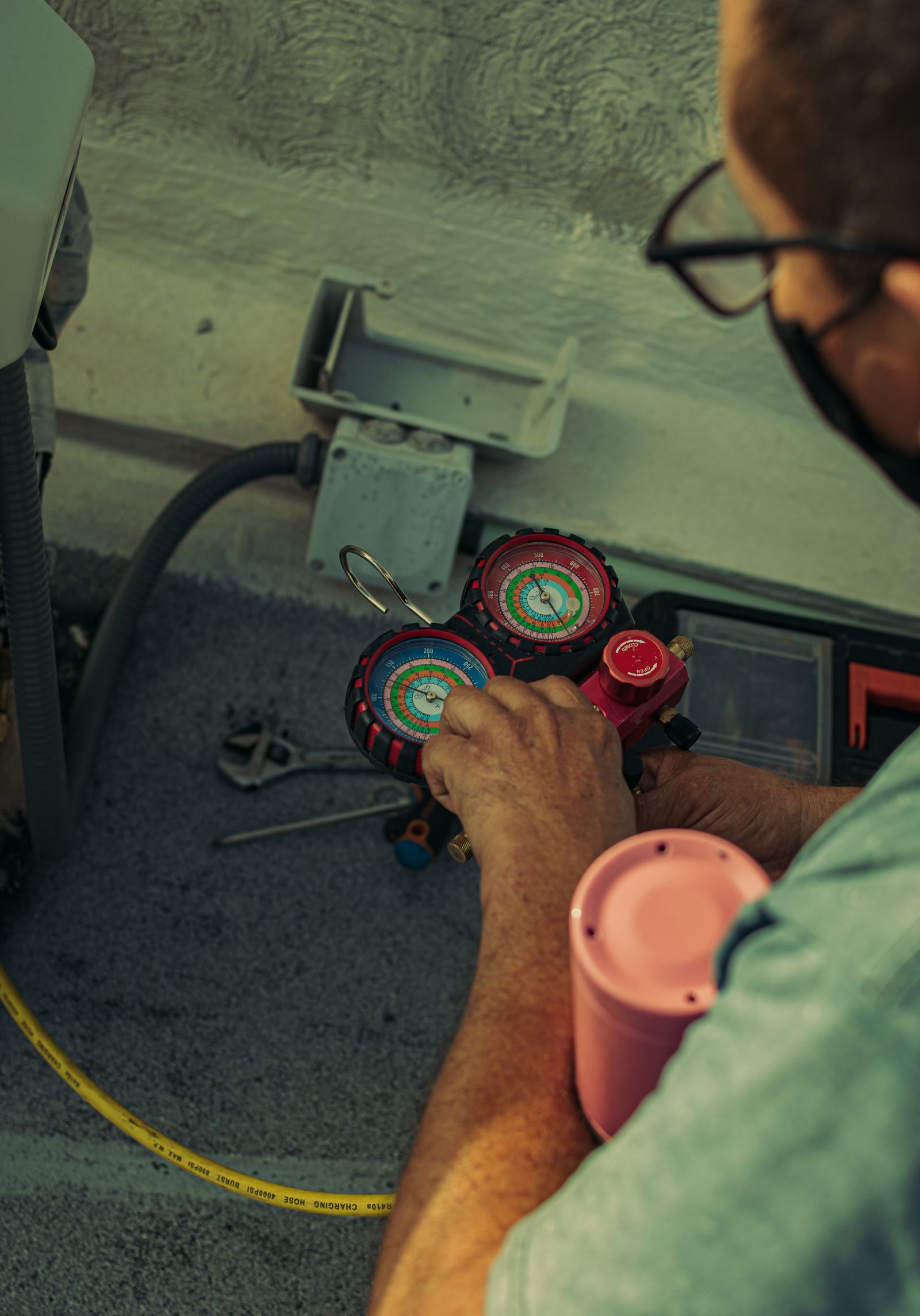 A technician holding a red-and-blue refrigerant manifold gauge set connected by a yellow charging hose to the internals of an outdoor air-conditioning unit, with hand tools on the ground.