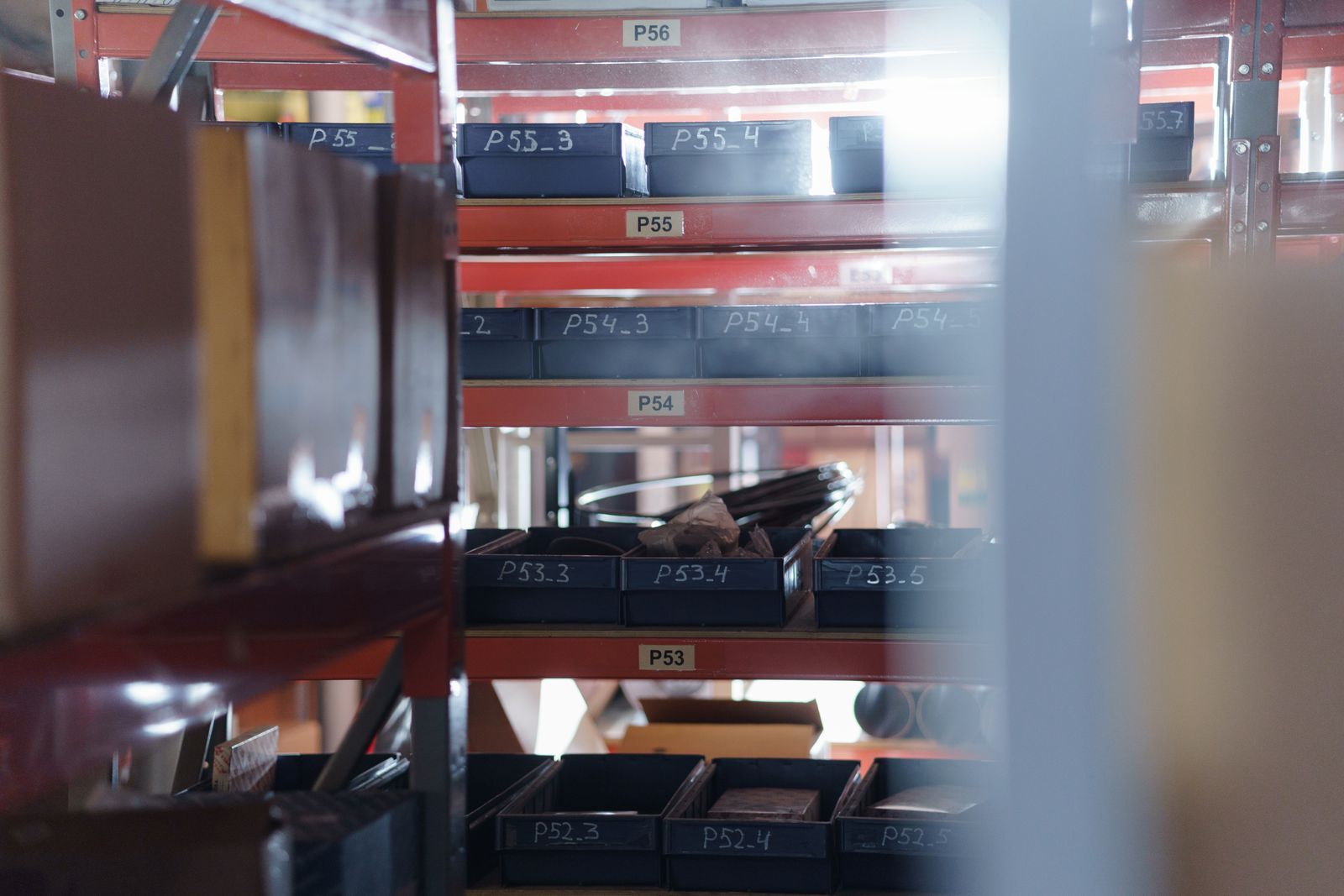 Rows of red industrial racking holding labelled black storage bins full of small parts and components in a storeroom.